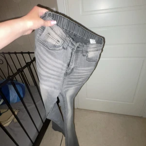 Grå bootcut jeans med resårmidja - Snygga grå jeans med bootcut-ben och resår i midjan för extra komfort. Klassisk femficksmodell med dragkedja och bälteshällor. Jeansen har en tvättad look och är perfekta för dig som gillar en avslappnad men trendig stil.