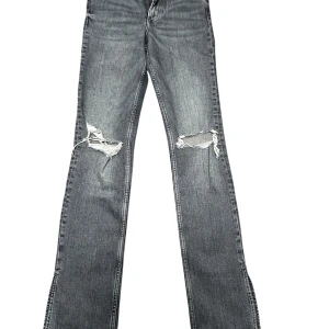 Gråa bootcut jeans med slits från Zara - Grå jeans från Zara med bootcut passform och slitna detaljer över knäna. Jeansen har klassisk femficksdesign, dragkedja och knapp framtill. Materialet är denim i bomull och färgen är tvättad grå med snyggt sliten look.