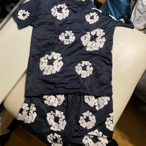 Mörkblå Denim Tears t-shirt - Säljer en mörkblå t-shirt från Denim Tears i storlek M. T-shirten har coola vita blommönster tryckta över hela framsidan och ärmarna. Rund hals och korta ärmar, tillverkad i mjuk bomull. Perfekt för dig som gillar streetwear och unika prints. Priset kan diskuteras och dom kan säljas som separata plagg. Alltså shortsen för sig och T-shirten för sig. Med vänlig hälsningar!