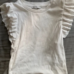 Vit ribbad topp med volangärm Zara - Superfin vit ribbad topp från Zara med korta volangärmar som ger en extra snygg touch. Mjuk och stretchig bomullskänsla, perfekt till jeans eller kjol. Enkel rund halsringning och figurnära passform. Säljes i storlek M. Använd ca 1-2 gånger.