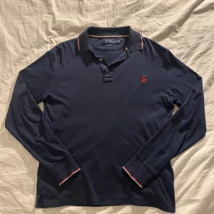 Marinblå långärmad piké från Polo Ralph Lauren - Snygg marinblå långärmad pikétröja från Polo Ralph Lauren i custom slim fit. Tröjan har klassisk krage med röd och vit kant, knappar vid halsen och ikonisk röd broderad logga på bröstet. Muddarna har matchande röd och vit detalj. 