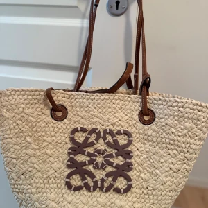 Beige shoppingväska från Loewe - Säljer en beige shoppingväska från Loewe med bruna läderhandtag och ett stort brunt Loewe-emblem framtill. Väskan är flätad i naturmaterial och har en rymlig form, perfekt för att bära med sig allt man behöver. Snygg och trendig detalj till din outfit.