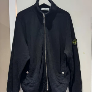 Svart zipjacka från Stone Island - Svart jacka från Stone Island med klassisk patch på ärmen. Jackan har hel dragkedja framtill, ståkrage och två fickor med knappstängning. Tillverkad i mjuk bomull för en clean och stilren look. Perfekt för dig som gillar streetwear och snygga detaljer.