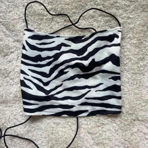 Zebramönstrad bandeau topp  - Snygg bandeau topp med  svartvita zebraränder och tunna svarta snören som kan knytas på olika sätt. Toppen är axelbandslös och har en croppad passform, perfekt för att sticka ut med ett djuriskt mönster. Materialet är mjukt och stretchigt polyester.