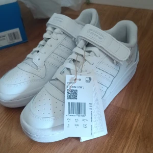 Helt nya Adidas Forum Low 00's  - Fräscha Adidas Forum Low J sneakers i helvit färg med klassiska tre ränder på sidan, snörning och kardborreband över vristen. Snygg retrostil med perforerad tå och platt sula. Tillverkade i skinn för en clean och sportig look. Perfekt för dig som gillar streetwear och sneakers. Säljer för att jag råkade beställa två par och de går inte att returnera!