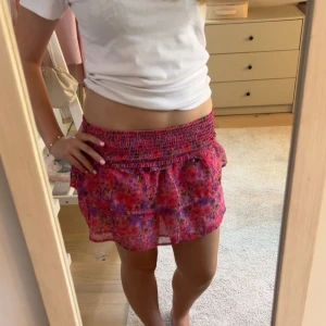 Blommig rosa volangkjol - Supersöt kort kjol med blommigt mönster i rosa, lila och röda toner. Kjolen har bred smock i midjan och dubbla volanger som ger en flowy känsla. Perfekt för dig som gillar färg och vill sticka ut lite extra i sommar.