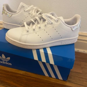 Adidas Stan Smith vita sneakers strl 36 - Fräscha vita Adidas Stan Smith sneakers med holografisk detalj på hälen som ger extra glans. Klassisk låg modell med snörning och perforerade ränder på sidorna. Skorna har rund tå och platt sula, tillverkade i syntetmaterial med mjukt foder för skön komfort.