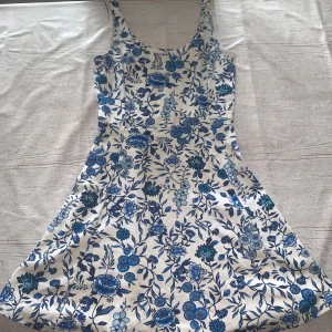 Vit och blå blommig sommarklänning - Supersöt kort klänning med blommigt mönster i blått på vit botten. Klänningen har ärmlös design och rundad hals, perfekt för varma dagar. Materialet känns lätt och luftigt, och den har en skön passform som ger en fin siluett.
