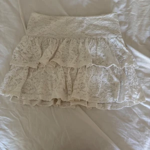 Vit spetskjol med volanger - Supersöt vit kortkjol med romantiska volanger och spetsdetaljer. Kjolen har flera lager och ett brett midjeband som ger en snygg siluett. Perfekt för dig som gillar en feminin och ljus stil.