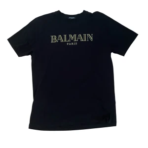 Svart Balmain Paris t-shirt i bomull - Svart t-shirt från Balmain Paris med klassisk rund hals och gul logotyptryck framtill. Tillverkad i 100% bomull för en skön och mjuk känsla. Enkel och stilren design som passar perfekt till jeans eller shorts.
