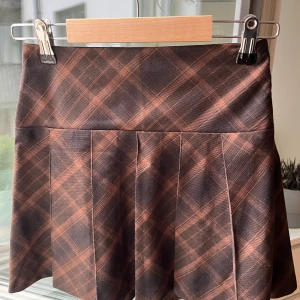 Brun rutig kortkjol från H&M Divided - Supersnygg brunrutig kortkjol från H&M Divided i storlek 32. Kjolen har klassiska veck och är gjord i mjuk polyester med lite stretch för skön passform. Perfekt för dig som gillar preppy vibes och vill sticka ut med ett coolt mönster.