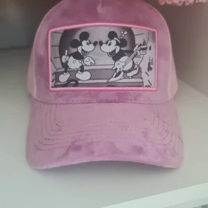 Rosa keps med Mickey & Minnie Mouse - Snygg rosa keps med mjuk sammet framtill och mesh baktill. Framsidan har en svartvit patch med Mickey och Minnie Mouse i retrostil. Justerbar snapback för perfekt passform. Perfekt accessoar för dig som gillar Disney och vill sticka ut. 