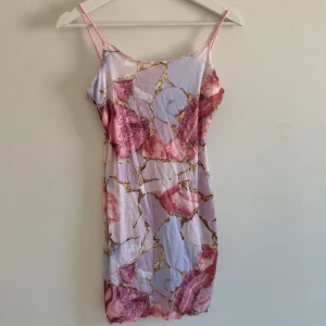 Pastellfärgad kort klänning från SHEIN - Säljer en kort klänning från SHEIN med tunna axelband och ett unikt abstrakt mönster i ljuslila, rosa och guld. Klänningen är figurnära och gjord i mjuk polyester med lite stretch. Perfekt för dig som gillar färg och vill sticka ut med en cool look.Kort Bodycon klänning med marmorerat mönster i guld, rosa och lila. Aldrig använd.