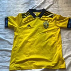 Svenska landslagströja Adidas gul - Gul fotbollströja från Adidas med Sveriges landslagsemblem på bröstet och mörkblå detaljer vid krage, axlar och ärmslut. Tröjan har klassisk V-ringning, korta ärmar och är tillverkad i lätt och ventilerande AEROREADY-material.