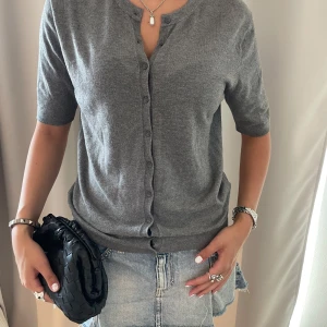 Grå kofta med kort ärm och knappar - Snygg grå kofta med korta ärmar och rund hals. Tröjan har knappar hela vägen framtill och är gjord i mjukt stickat material. 