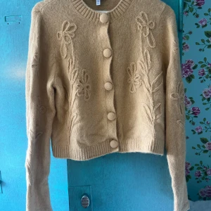 Beige kofta från & Other Stories - Superfin beige kofta från & Other Stories med stora, matchande knappar och broderade blommor framtill och på ärmarna. Koftan är stickad i mjukt material och har långärmad passform med ribbad kant vid hals och nederdel. Helt ny men saknar prislapp. Aldrig använd.