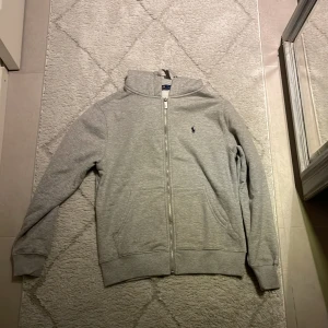 Grå zip från Polo Ralph Lauren - Säljer en sprillans ny stilren grå hoodie från Polo Ralph Lauren med dragkedja och klassisk logga på bröstet. Tröjan har huva med snörning, två fickor fram och ribbade muddar vid ärmslut och nederkant. Perfekt för chill dagar eller när du vill ha en clean look. Om du är intresserad är det bara att föreslå pris!