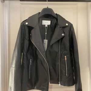 Ny svart skinnjacka från Vila - Helt ny snygg, svart skinnjacka från Vila med klassisk bikerstil. Jackan har silvriga dragkedjor, bred krage och två fickor framtill. Materialet är slätt och jackan har lång ärm. Perfekt för dig som vill ha en edgy look.