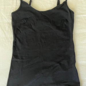 Svart linnetopp med smala axelband - Enkel och stilren svart linnetopp med smala justerbara axelband. Toppen har en rak passform och är tillverkad i mjukt bomullsmaterial, perfekt att matcha med jeans eller kjol. Snygg bas till garderoben för en clean look.