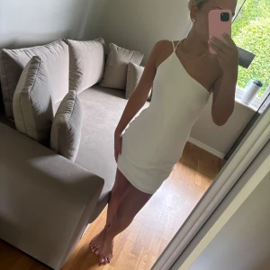 Vit kort klänning med smala axelband - Säljer en vit kort klänning med smala axelband och oneshoulder-design. Klänningen har en enkel och stilren look med figurnära passform och öppen rygg. Perfekt för varma dagar och snygg att styla med smycken eller sandaler.