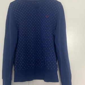 Blå prickig tröja från Fred Perry - Mörkblå långärmad tröja från Fred Perry med små vita prickar över hela framsidan och baksidan. Klassisk rund halsringning och diskret Fred Perry-logga broderad på bröstet. Skön bomullskvalitet som passar perfekt till jeans eller kjol.