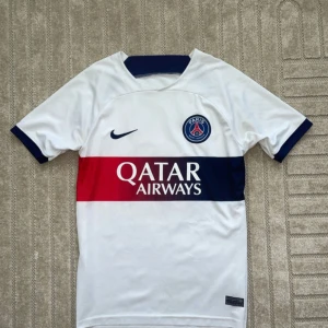 PSG Mbappé vit matchtröja Nike - Säljer en officiell Paris Saint-Germain fotbollströja med Mbappé och nummer 7 på ryggen. Tröjan är vit med breda blå och röda ränder över bröstet, PSG-emblem på vänster sida och Nike-logga på höger. Kortärmad och tillverkad i lätt polyester, perfekt för träning eller match.