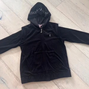 Svart velourjacka Juicy Couture - Svart velourjacka från Juicy Couture med huva och dragkedja. Stora Juicy Couture-stenar på huvan och JC-logga på bröstet. Jackan har känguruficka och är långärmad, perfekt för en chill och trendig look.