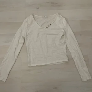 Vit långärmad tröja från Mango - Vit långärmad tröja från Mango med v-ringad hals och tre små dekorativa knappar vid halsringningen. Tröjan är i mjuk bomull och har en enkel, clean look som funkar till allt. Perfekt för dig som gillar minimalistisk stil och vill ha något bekvämt. Knappt använd ca 2 gånger.