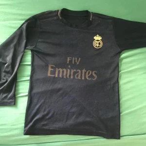 Real Madrid Hazard #7 långärmad tröja - Snygg svart långärmad Real Madrid fotbollströja från Adidas med guldiga detaljer och tryck. På framsidan finns klubbmärket och Fly Emirates-loggan, på ryggen står Hazard och nummer 7. Materialet är lätt och andas, perfekt för dig som gillar fotboll och vill ha en cool look.