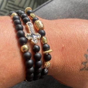 Svarta pärlarmband med gulddetaljer - Tre snygga armband med mattsvarta pärlor och guldfärgade detaljer. Ett av armbanden har ett coolt kors med glittrande stenar, medan de andra har olika mönstrade och släta guldfärgade pärlor. Perfekt för att ge din stil lite edge och bling.