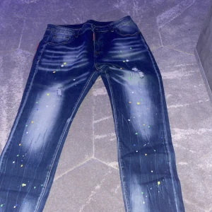 Dsquared2 jeans med färgstänk - Snygga blå jeans från Dsquared2 med coola färgstänk i flera färger och slitna detaljer. Baktill finns stora patchar och logga. Jeansen har en slim passform och är tillverkade i bomull. Perfekta för dig som vill sticka ut med din stil.