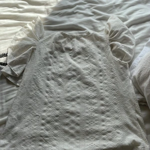 Vit blus med puffärmar och broderi - Supersöt vit blus med puffiga korta ärmar och broderat mönster över hela tyget. Blusen har rund halsringning och romantisk känsla, perfekt för dig som gillar detaljer och en feminin stil. Tyget är lätt och luftigt, vilket gör den skön att bära under varmare dagar.