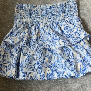 Blåvit volangkjol från Gina Tricot - Supersöt kort kjol från Gina Tricot med blått och vitt paisleyinspirerat mönster. Kjolen har bred smock i midjan och dubbla volanger som ger en skön och flowig känsla. Perfekt för dig som gillar färg och mönster!