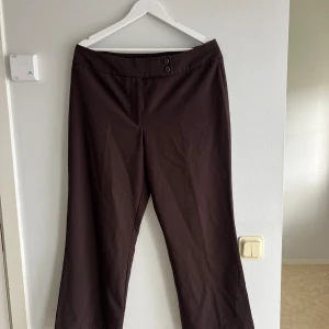 Bruna kostymbyxor från Haggar - Mörkbruna kostymbyxor från Haggar med klassisk rak passform och två knappar i midjan. Byxorna har en stilren look och är tillverkade i ett mjukt bomullsmaterial. Perfekta för dig som gillar en enkel och tidlös stil.