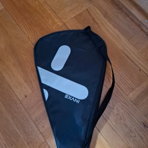 Svart racketväska från Exani - Säljer en svart racketväska från Exani med vitt tryck. Väskan har en axelrem och är formad för att passa ett racket, perfekt för att bära till och från träningen. Enkel och stilren design.