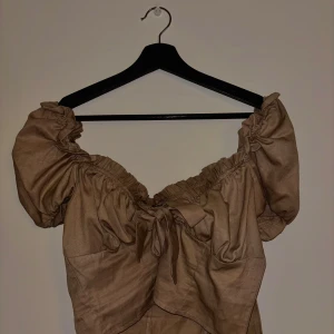 Beige offshoulder blus från NA-KD - Säljer en beige offshoulder blus från NA-KD med puffiga ärmar och knytning framtill. Blusen har en romantisk och trendig look med volangdetaljer vid ärmslut och halsringning. Perfekt för att skapa en chic outfit.