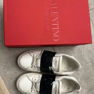 Valentino Garavani sneakers med mörkblå detalj - Säljer ett par vita sneakers från Valentino Garavani med bred svart rem över vristen och klassisk logga på plösen. Skorna har rund tå, snörning och platt sula. Kommer med originalkartong. Något nötta insida häl, inget som syns när man bär dem.