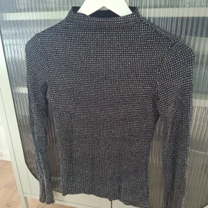 Glittrig svart långärmad topp från H&M - Säljer en svart långärmad topp från H&M med glittrigt mönster i flera färger. Toppen är tight och har en hög hals, perfekt för att sticka ut lite extra. Superstretchigt material som sitter snyggt på kroppen.