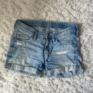 Ljusblå jeansshorts från &Denim - Säljer ett par ljusblå jeansshorts från &Denim med klassisk femficksdesign och uppvikta benslut. Shortsen har slitna detaljer framtill och en liten läderpatch bak i midjan. Perfekta för varma dagar.
