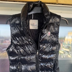 Svart dunväst från Moncler - Snygg svart dunväst från Moncler med hög krage och dragkedja framtill. Västen har Moncler-logga på bröstet och en glansig finish. Perfekt för lager-på-lager och håller dig varm under kyliga dagar.