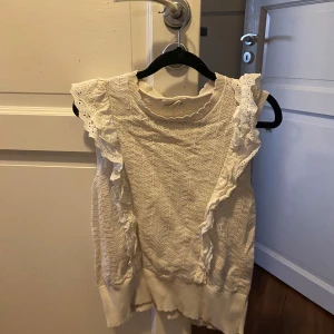 Vit stickad ärmlös topp med spetsdetaljer - Supersöt vit stickad topp med ärmlös design och breda axelpartier dekorerade med spetsvolanger. Snyggt mönster i stickningen och rund halsringning. Perfekt att styla till jeans eller kjol för en fräsch look.