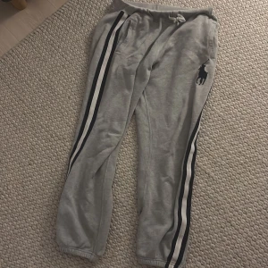 Grå mjukisbyxor med svarta och vita ränder från Polo Ralph Lauren - Säljer ett par grå mjukisbyxor från Polo Ralph Lauren med svarta och vita ränder längs sidorna. Byxorna har resår och snörning i midjan samt elastiska benslut. Perfekta för en avslappnad stil.