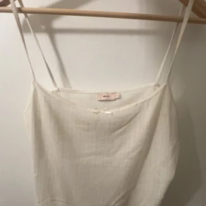 Vit linnetopp från Nelly - Säljer en söt vit linnetopp från Nelly med tunna axelband och diskret strukturmönster. Toppen har en liten rosett framtill och är perfekt för varma dagar. Enkel och stilren design som passar till mycket.