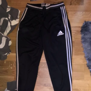 Svarta Adidas träningsbyxor med vita ränder - Säljer ett par klassiska adidas byxor i ett syntetmaterial som passar perfekt för träning. med Adidas-loggan på låret. Byxorna har dragkedja längst ner på benen ifall man inte vill ha dom tajt, även fickor med dragkedja på sidan av låren. Dem är helt nya, säljer dem då dem aldrig kommer till användning 