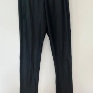 Svarta leggings från Name It - Svarta fejk skinn leggings från Name It i stretchigt material. De har en enkel och stilren design med resår i midjan, perfekta för en bekväm och snygg look.