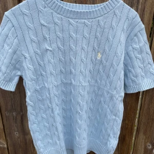 Ljusblå kabelstickad tröja från Polo Ralph Lauren - Säljer en ljusblå kabelstickad tröja från Polo Ralph Lauren med korta ärmar och rund hals. Tröjan har det klassiska broderade logotypen på bröstet och är gjord i mjukt material. Perfekt för en stilren och avslappnad look. unisex och kan passa man xs/s