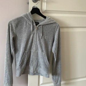 Grå hoodie med dragkedja från Juicy Couture - Säljer en klassisk grå hoodie från Juicy Couture i mjukt material. Tröjan har dragkedja framtill, huva med snören och två fickor. Perfekt för en avslappnad och stilren look.