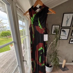 Svart långklänning med grönt och rött mönster från Rodebjer - Säljer en svart långklänning från Rodebjer med stora gröna och röda mönster. Klänningen har omlott-detalj upptill och breda axelband samt fickor. Tyget är mjukt och faller snyggt, perfekt för dig som vill sticka ut. Aldrig använd. Nypris 4995 kr. Säljes för 900 kr