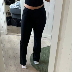 Svarta kostymbyxor med bootcut - Säljer ett par svarta kostymbyxor med bootcut passform och hög midja. Byxorna har klassisk look med raka ben som blir lite utsvängda nertill. Perfekta till en snygg topp eller skjorta. Den har en snygg metall detalj som är ett ”J” på sidan. 