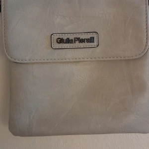 Ljusgrå axelväska från Giulia Pieralli - Säljer en stilren ljusgrå axelväska från Giulia Pieralli. Väskan har ett lock framtill med logotyp, justerbar axelrem och dragkedjefack på baksidan. Perfekt storlek för det viktigaste och enkel att matcha till olika outfits.